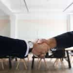 Two hands shaking in front of a long table in a meeting room inside of an office.