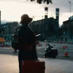 A traveler stands in a town square holding a red suitcase, backpack and hat