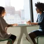 Two women sitting at a small table in an office environment while speaking to one another.